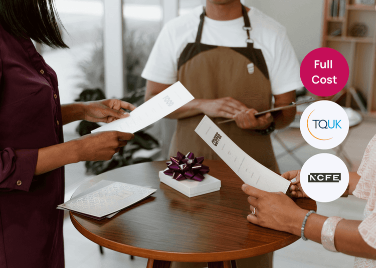 Two customers reading menus at a small table while a staff member in an apron stands nearby in a hospitality setting.