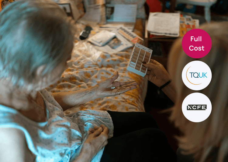 Older woman sitting on a bed holding tablets in her hand while a carer shows a medication chart for guidance.