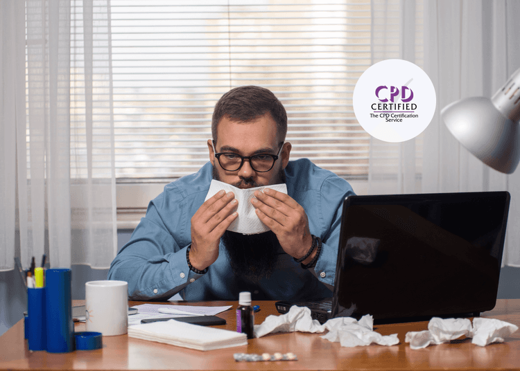 Man in glasses at a cluttered desk blowing his nose with tissues and medicine bottles in front of him.