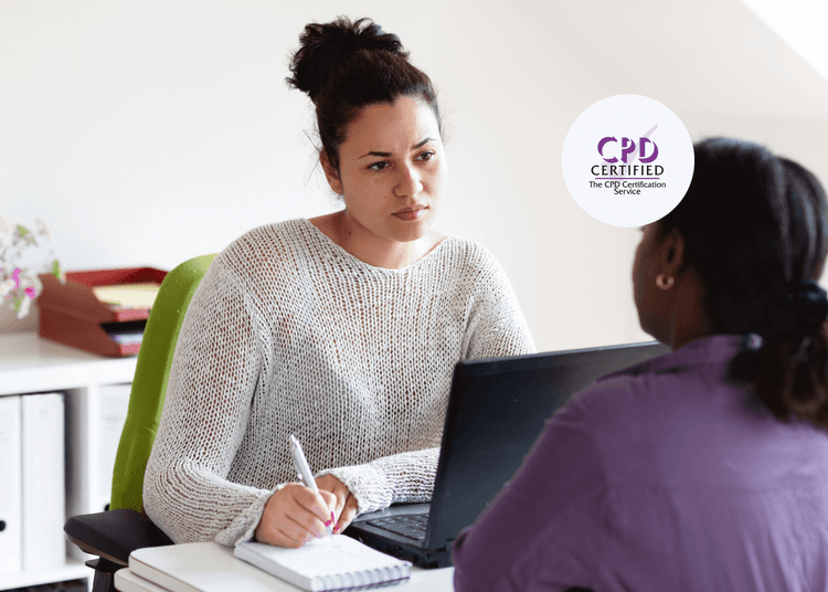 Woman in a white jumper listening intently and taking notes during a conversation across a desk with another person.