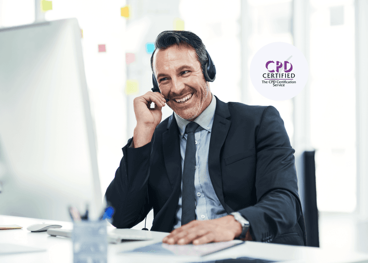 Smiling man in a suit wearing a headset, sitting at a desk with a computer and notepad.