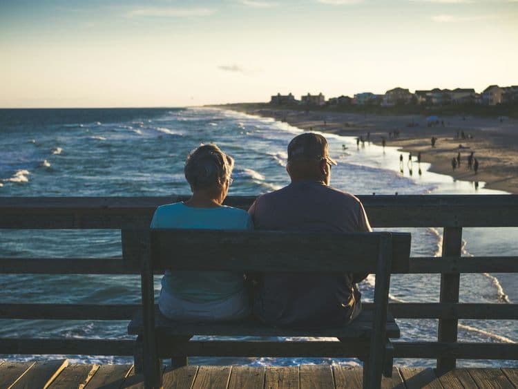 An older couple sit side by side on a wooden bench overlooking the sea from a pier, watching waves and people walking along the shoreline at sunset.