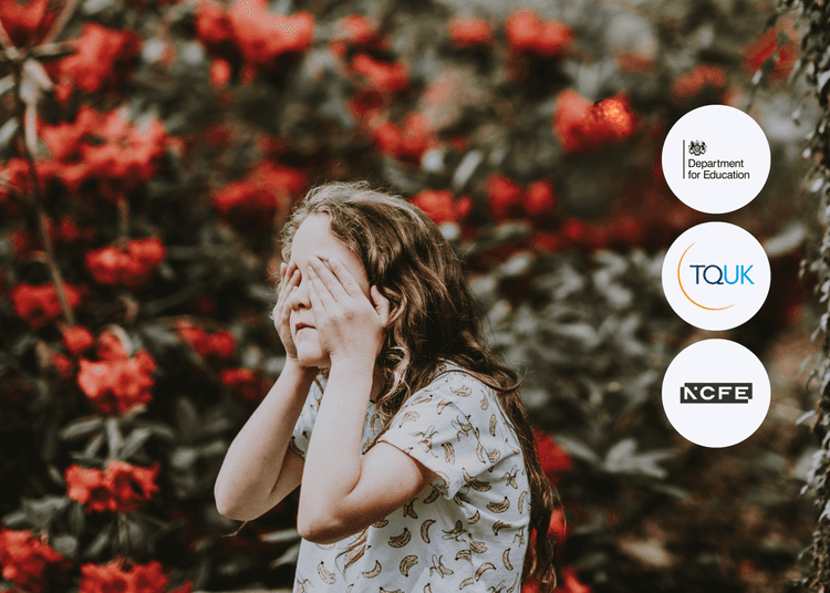 Child with long hair covers their face with both hands, standing in front of red flowering bushes.
