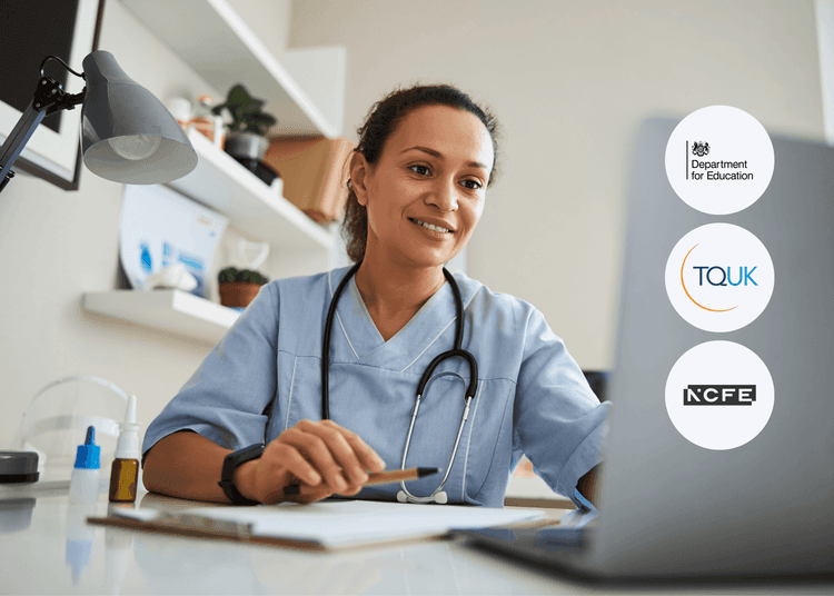Smiling healthcare worker in scrubs with a stethoscope sits at a desk, using a laptop beside a notepad and medicine bottles.