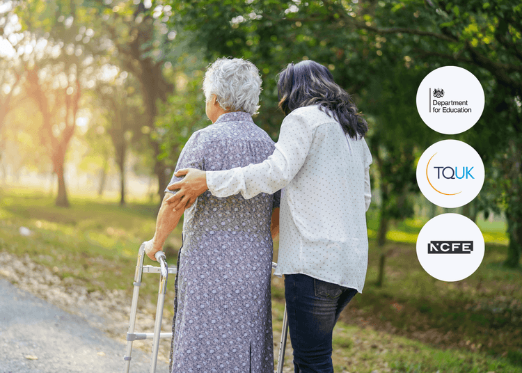 Caregiver supporting an older woman using a walking frame along a park path lined with trees on a sunny day.