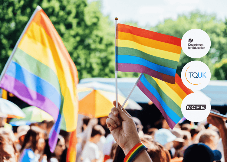 Hand holding a small rainbow flag at an outdoor pride event with people, umbrellas, and trees visible in the background.
