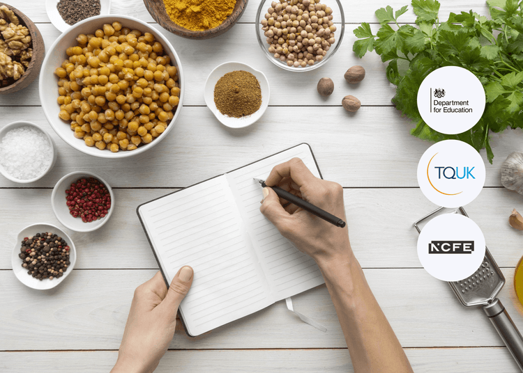Hands holding an open lined notebook beside bowls of chickpeas, spices, herbs, and peppercorns on a white wooden surface.
