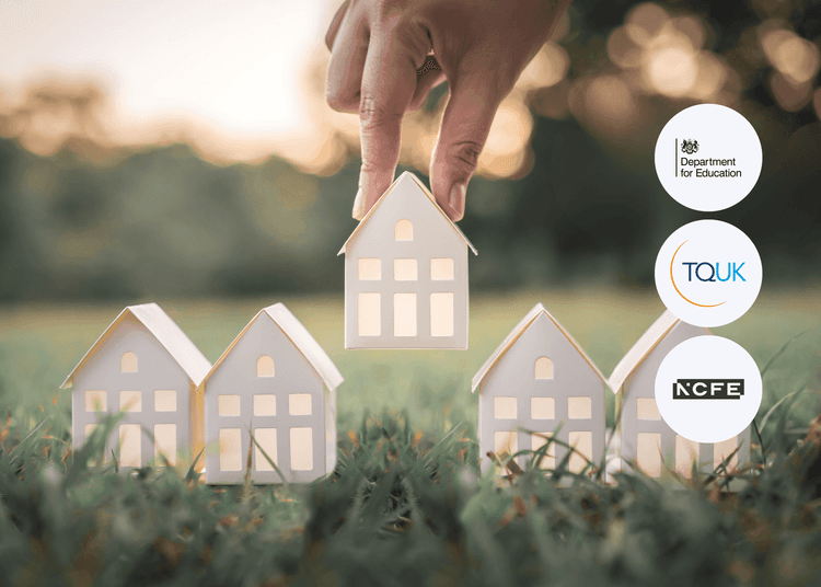 A hand lifting a small white model house from a row of identical houses placed on grass, with trees in the background.