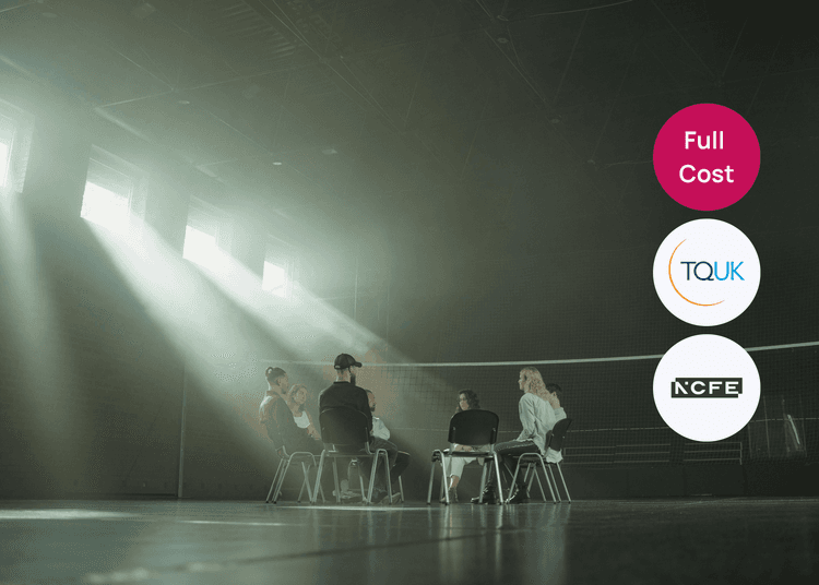 A small group sits together in a circle in a bright hall, reflecting a supportive setting for mental health training. Group of people seated in a circle in a large hall with sunlight streaming through high windows during a support session.