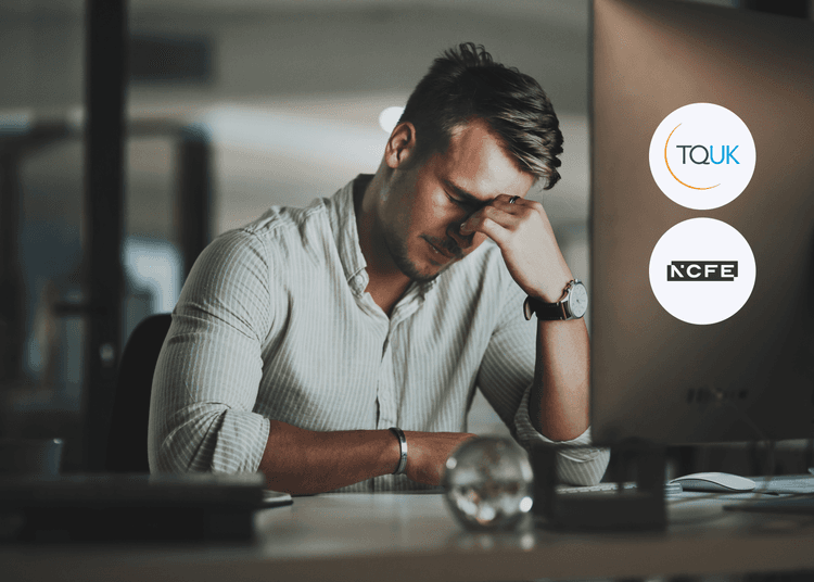 A man showing signs of stress while working at his desk, illustrating the emotional challenges addressed in the Behaviour that Challenges course. A stressed man sitting at a desk with his head in his hand beside a computer displaying TQUK and NCFE logos, representing workplace pressure.