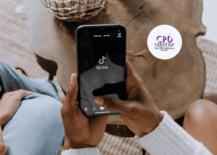 A TikTok launch screen shown on a mobile device, supporting CPD learning on youth engagement with social media platforms.  Close-up of hands holding a smartphone displaying the TikTok app home screen, with two people seated on a wooden table.