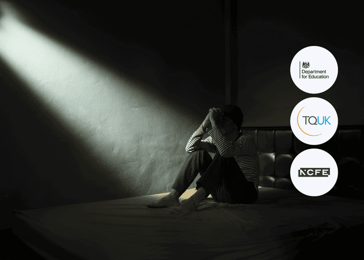 A person sits alone on a bed under dim lighting, used to reflect themes in suicide awareness and self-harm prevention. Person sitting on a bed in a dark room with their head in their hands, lit by a narrow beam of light coming through the wall.