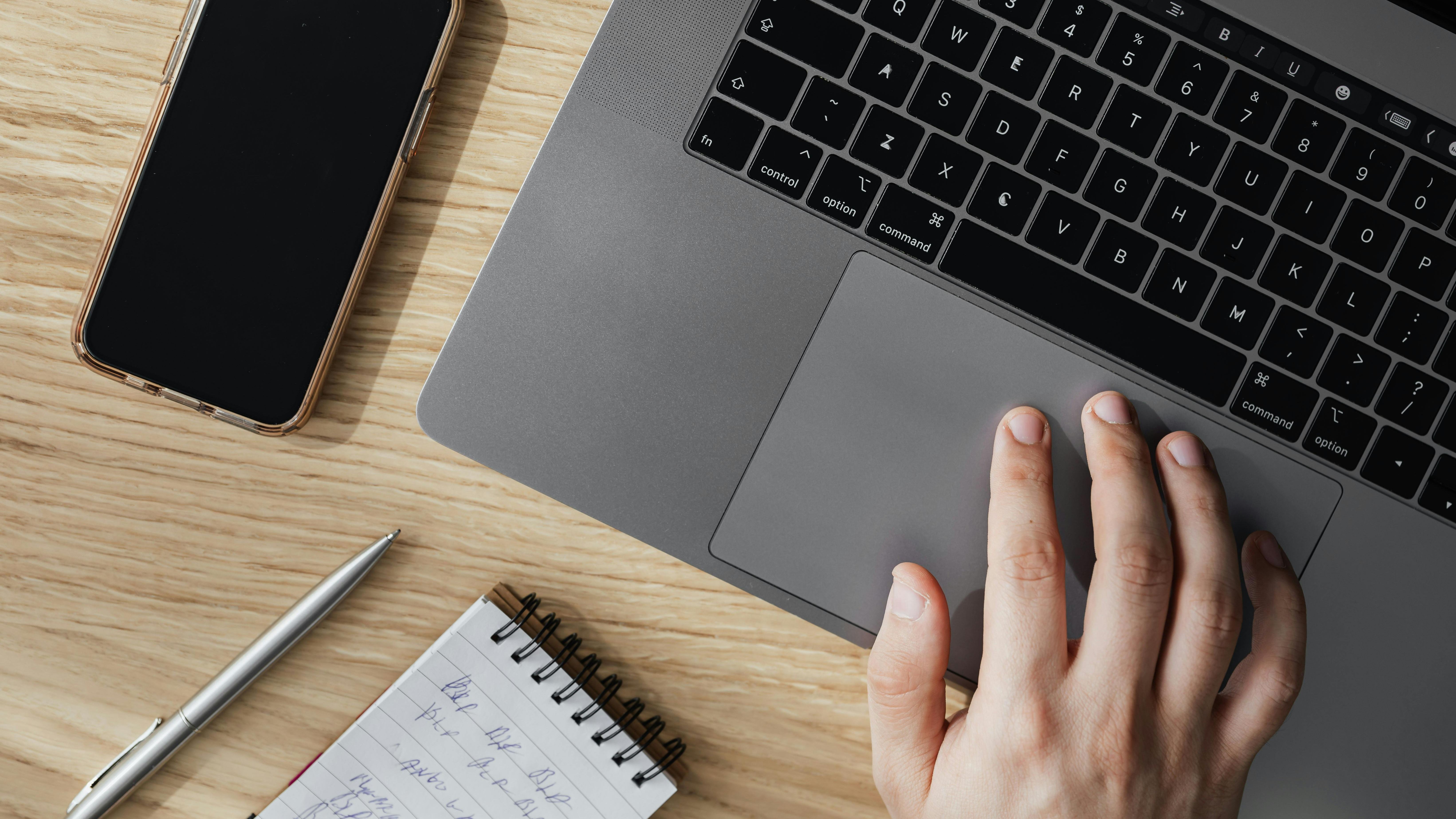 A mans hand hovering over a grey laptop. Next to the laptop is a phone, notepad, and pen